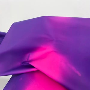Puede incluir: Un trozo de tela con un degradado de color. La tela pasa de un morado intenso a un rosa vibrante. El material es liso y parece estar doblado, creando sombras y reflejos en su superficie.