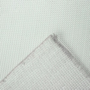 May include: Close-up of a light gray fabric with a woven texture. The corner of the fabric is folded over, revealing the edge. The fabric appears to be a light, neutral color.