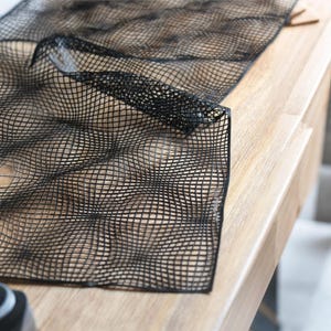Puede incluir: Una hoja de malla negra flexible con un patrón de cuadrícula repetitivo de líneas que se cruzan. El material es fino y semitransparente, mostrando un diseño geométrico que crea una sutil ilusión óptica de profundidad en la superficie.