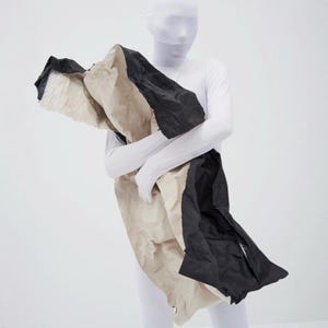 Op de afbeelding: Een grote, abstracte sjaal in zwart, beige en wit. De sjaal lijkt gemaakt van gelaagde stof met een gekreukte textuur. Het ontwerp is gedrapeerd en wordt vastgehouden voor een persoon die een wit bodysuit draagt.