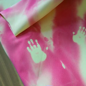 以下が含まれることがあります： ピンクと黄色のタイダイ効果のある生地。生地には、薄い緑色の手形模様があります。生地は折りたたまれており、裏面は無地の黄色です。