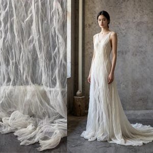 Puede incluir: Un vestido blanco sin mangas hasta el suelo con escote en V y una capa de tela texturizada y arrugada que crea pliegues verticales que caen hasta el suelo, formando una cola que se extiende por el suelo en una silueta fluida.