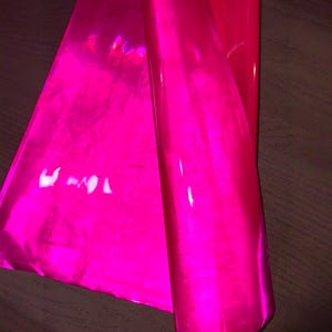 May include: A roll of vibrant, hot pink vinyl material. The material is glossy and reflects light, creating highlights and shadows. The vinyl is partially unrolled, revealing its flexible nature. The background is a dark wood surface.