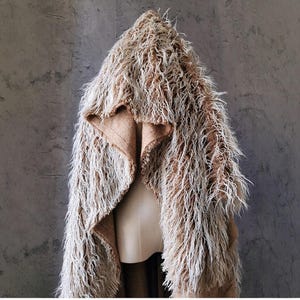 May include: A hooded jacket with a shaggy, textured appearance. The jacket features a mix of brown and off-white fibers, creating a layered, fur-like effect. The interior lining is a solid, light brown fabric. The jacket is displayed on a mannequin against a gray background.