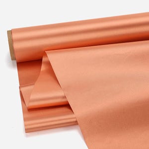 May include: A roll of peach-colored fabric with a satin finish. The fabric is partially unrolled, revealing the smooth texture and sheen. The roll is made of a cardboard core.