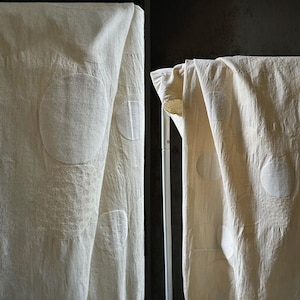 Puede incluir: Cortinas de tela blanquecina con grandes patrones circulares texturizados. Las cortinas están drapeadas sobre una barra, con pliegues y sombras suaves. La tela parece ser lino o un material natural similar, creando una estética simple y elegante.