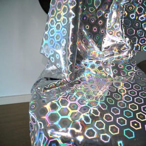 May include: Silver holographic fabric with a repeating honeycomb pattern. The fabric reflects a rainbow of colors, creating a shimmering effect. The material is draped, showing its texture and flexibility. Ideal for crafts and decor.