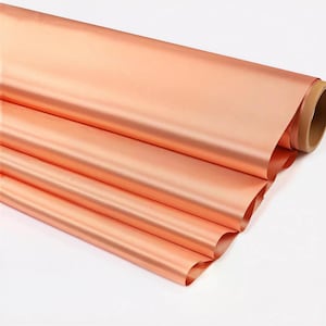 May include: A roll of copper-colored metallic foil. The foil is a warm, reddish-orange hue with a smooth, reflective surface. The roll is partially unraveled, revealing several layers of the foil.