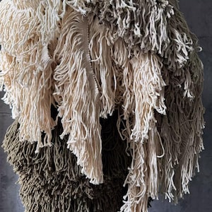 May include: Close-up of a textured textile in cream, tan, and olive green. The fabric is made of thick, knotted yarn or rope, creating a fringed effect. The layers overlap, adding depth and dimension. The textile is likely for home decor.