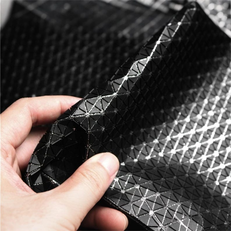 May include: A flexible black material featuring a repeating geometric pattern of small raised triangles. The surface is composed of interconnected black triangular segments with thin, reflective silver lines running between each individual shape.