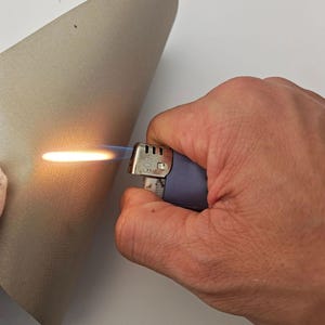 May include: A blue and silver lighter with a flame ignited, held by a person. The flame is orange and blue, illuminating the edge of a beige material. The lighter's metal top has a series of small holes.
