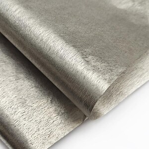 May include: Close-up of a folded piece of gray faux fur fabric. The fabric has a soft, textured appearance with a slight sheen. The folds create shadows and highlights, adding depth to the material. The fabric appears to be a light gray color.