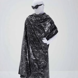 Puede incluir: Un chal con patrón de mármol negro y plateado sobre un traje blanco de cuerpo completo. El chal presenta un acabado metálico de alto brillo con vetas blancas intrincadas. El conjunto incluye una cubierta para la cabeza blanca y gafas oscuras.