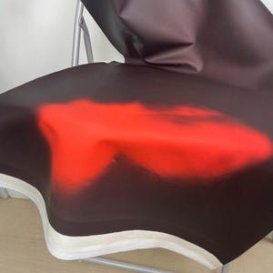 May include: A folding chair with a black seat featuring a large, abstract red design. The seat has a white border and is supported by a silver metal frame. The chair is designed for indoor or outdoor use.