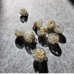 May include: A collection of clear glass orb earrings with gold-colored metal findings. Each orb contains small, dried, beige-colored flowers and delicate silver-colored wire. The earrings are displayed on a gray surface.
