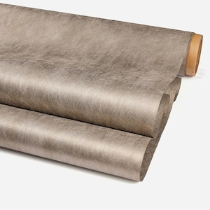 May include: A roll of taupe-colored fabric with a textured, distressed appearance. The fabric is rolled up, revealing the edge of the roll, and partially unrolled, showing the material's surface. The fabric has a subtle sheen.