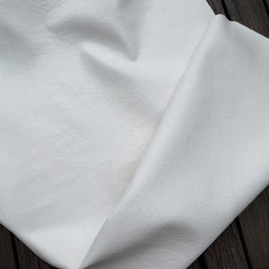 Puede incluir: Primer plano de una tela blanca texturizada con un aspecto ligeramente arrugado. El material está doblado, revelando el reverso. La tela tiene una sutil veta similar al cuero.