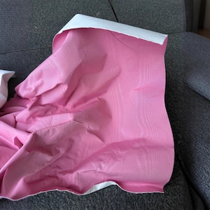 May include: A piece of fabric featuring a vibrant pink top surface with a subtle, fine-grained texture and a solid white underside. The material is draped to show folds and creases, highlighting the contrast between the pink and white layers.