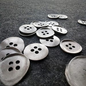 Peut inclure: Plusieurs boutons argentés, chacun percé de quatre trous, disposés sur une surface gris foncé. Les boutons sont ronds et présentent un aspect légèrement texturé, suggérant un matériau métallique.