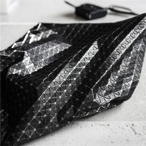 May include: A black bag featuring a geometric, triangular grid pattern across its surface. The material has a glossy, reflective finish that catches the light, creating a textured, faceted appearance with sharp angles and a structured, folded form.