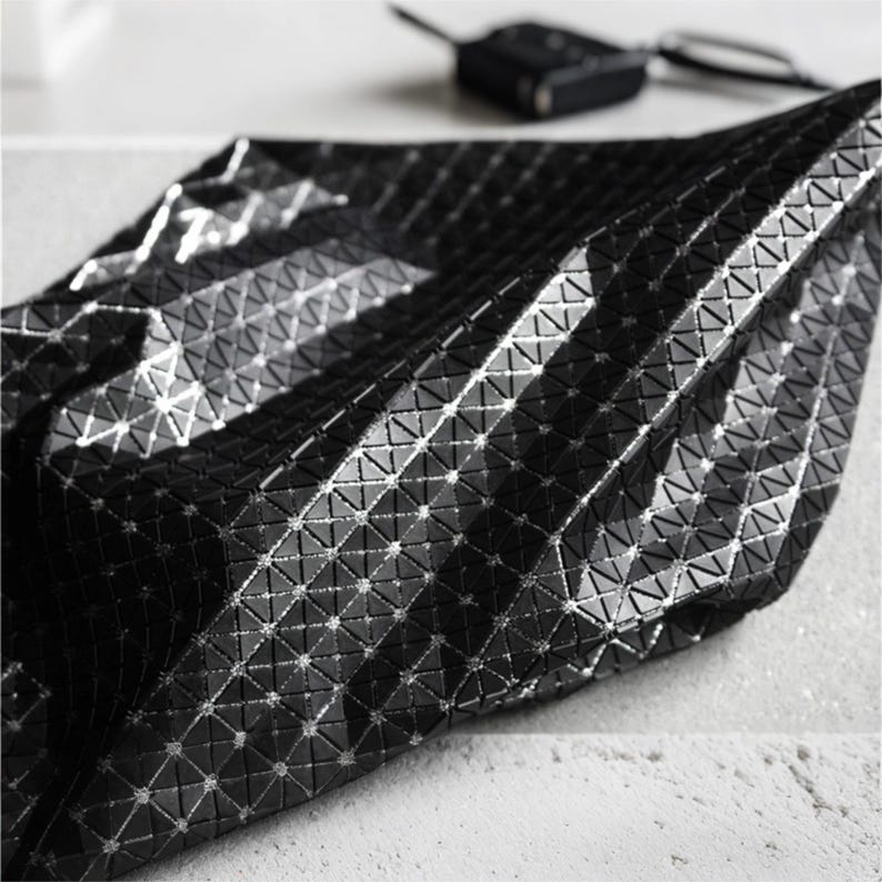 May include: A black bag featuring a geometric, triangular grid pattern across its surface. The material has a glossy, reflective finish that catches the light, creating a textured, faceted appearance with sharp angles and a structured, folded form.