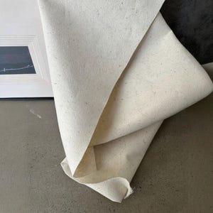 May include: Off-white, textured fabric folded over, revealing layers and a rolled edge. The fabric has a speckled appearance and is positioned near a framed artwork with a minimalist design. The overall aesthetic is neutral and natural.