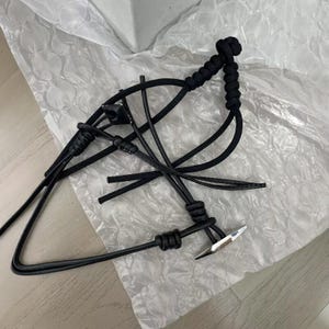 May include: A black cord necklace featuring multiple decorative knots along the length of the cord. The necklace is secured with a silver-toned metal toggle clasp, which consists of a T-bar and a circular ring component at the ends of the cord.