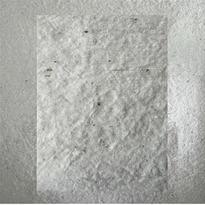 May include: Close-up of a textured white fabric with a slightly irregular surface. The fabric has a woven appearance with small, dark specks scattered across it. The texture suggests a natural fiber material.