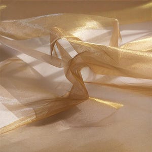 Puede incluir: Primer plano de una tela fluida y transparente en tonos dorados y beige. El material está delicadamente drapeado, creando pliegues y reflejos suaves. La tela tiene un brillo sutil, lo que sugiere una textura lujosa, y es ideal para fines decorativos.