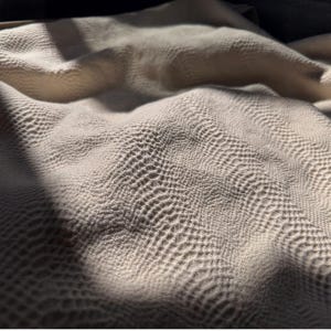 May include: Close-up of a textured, off-white fabric with a repeating pattern. The fabric has a subtle, rippled effect, with shadows adding depth and dimension. The overall appearance suggests a soft, luxurious textile.