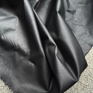 May include: A close-up of a black fabric with a subtle sheen. The material is folded and draped, revealing the texture and weave of the textile. The fabric appears smooth and has a slight sheen, suggesting it may be a synthetic material.