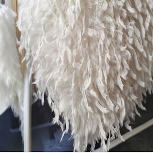 May include: Close-up of a white, feathery textile. The texture is dense and layered, with individual feathers visible. The overall effect is soft and voluminous, suggesting a decorative item or garment.