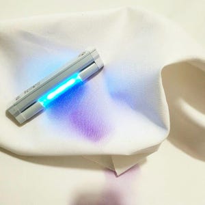 Puede incluir: Un bolígrafo quitamanchas blanco con una luz azul brillante que ilumina una tela blanca. Una mancha morada es visible en la tela. El bolígrafo es gris y rectangular.