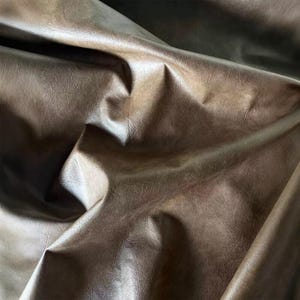 May include: A piece of brown faux leather fabric with a smooth, slightly distressed texture. The material is draped in deep folds, creating a series of highlights and shadows across the surface that emphasize the supple, flexible quality of the material.