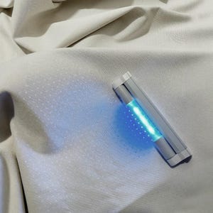 May include: A silver and gray UV light sanitizer with a bright blue light is shown on a light gray fabric. The sanitizer is rectangular with rounded edges. The fabric has a subtle dotted pattern.
