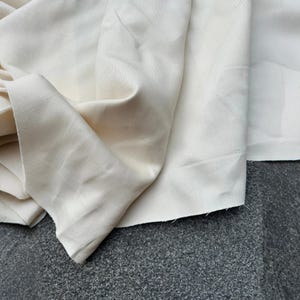 May include: A length of off-white, unbleached cotton canvas fabric featuring a visible twill weave texture. The material is draped in soft folds, showcasing its natural, matte finish and raw, slightly frayed edges along the cut bottom hem.