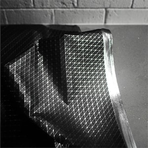 May include: A black fabric sheet featuring a repeating geometric pattern of small raised triangles and dots. The material has a reflective, metallic sheen that catches the light, creating highlights across the textured surface and folds.