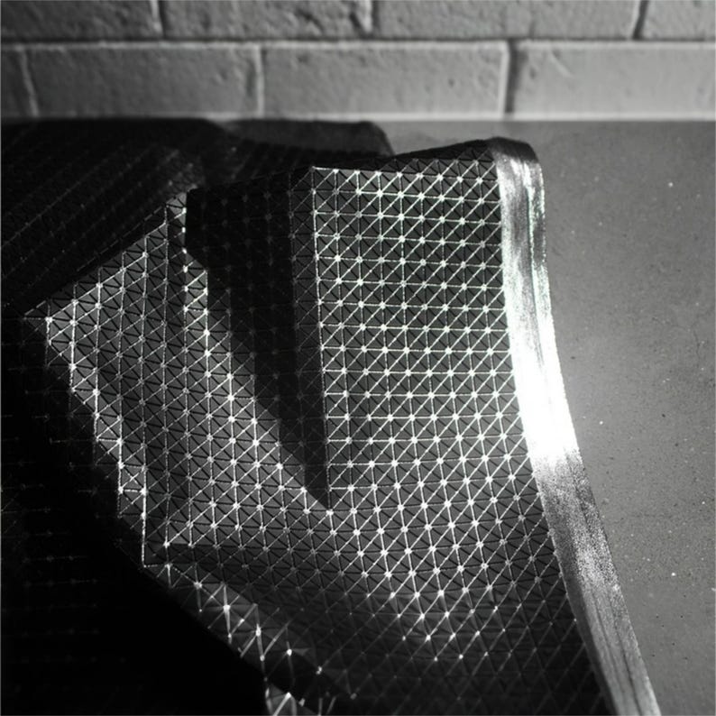May include: A black fabric sheet featuring a repeating geometric pattern of small raised triangles and dots. The material has a reflective, metallic sheen that catches the light, creating highlights across the textured surface and folds.