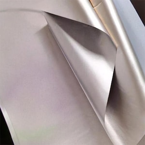 May include: A close-up of a sheet of pearlescent paper with a corner folded over, revealing a darker, metallic underside. The paper has a smooth, reflective surface, and the lighting creates highlights and shadows.