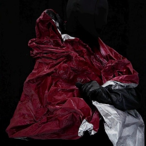 Op de afbeelding: Een gekreukeld rood, zwart en zilver materiaal tegen een zwarte achtergrond. Het rode materiaal lijkt een kledingstuk met capuchon te zijn. Het zwarte materiaal is een handschoen. Het zilveren materiaal is reflecterend.