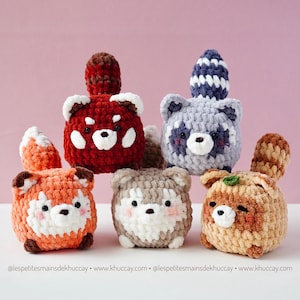 Pode incluir: Uma coleção de seis bichos de pelúcia de animais de crochê feitos à mão. Os bichos de pelúcia incluem um panda vermelho, um guaxinim, uma raposa, um husky e um urso. Cada bicho de pelúcia tem um design único com cores e características diferentes. O fundo é rosa liso.