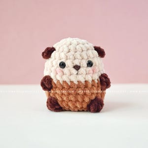 K&ouml;nnte beinhalten: Handgeh&auml;kelter Otter-Pl&uuml;sch in Creme, Braun und Dunkelbraun. Der Otter hat einen runden K&ouml;rper, kleine Ohren und ein s&uuml;&szlig;es Gesicht mit rosa Wangen und schwarzen Augen. Der Pl&uuml;sch liegt auf einer wei&szlig;en Oberfl&auml;che vor einem rosa Hintergrund.