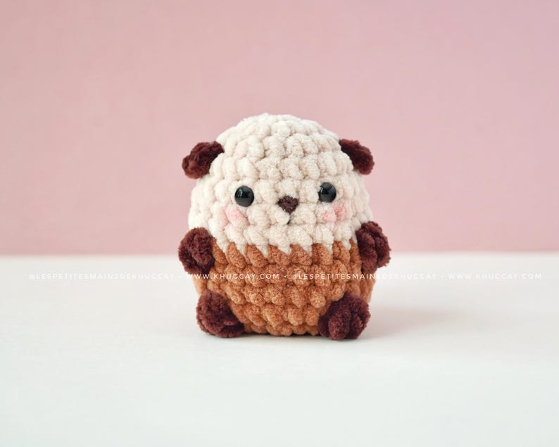 K&ouml;nnte beinhalten: Handgeh&auml;kelter Otter-Pl&uuml;sch in Creme, Braun und Dunkelbraun. Der Otter hat einen runden K&ouml;rper, kleine Ohren und ein s&uuml;&szlig;es Gesicht mit rosa Wangen und schwarzen Augen. Der Pl&uuml;sch liegt auf einer wei&szlig;en Oberfl&auml;che vor einem rosa Hintergrund.