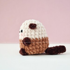 K&ouml;nnte beinhalten: Ein handgeh&auml;keltes Otter-Pl&uuml;schtier in Creme, Braun und Dunkelbraun. Das Stofftier hat einen abgerundeten K&ouml;rper, kleine Ohren und einen Schwanz. Das Gesicht des Otters hat kleine schwarze Augen und rosige Wangen.