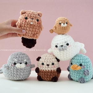 May include: A collection of handmade crochet animal plushies in various colors. The plushies include a beaver, a bird, a seal, a panda, a platypus, and a gray seal. The beaver is held by a hand. The background is a solid pink.