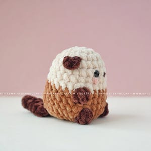 K&ouml;nnte beinhalten: Ein handgefertigtes geh&auml;keltes Otter-Pl&uuml;schtier in Creme, Braun und Dunkelbraun. Der Otter hat einen cremefarbenen Kopf mit dunkelbraunen Ohrakzenten, einen braunen K&ouml;rper und einen dunkelbraunen Schwanz.