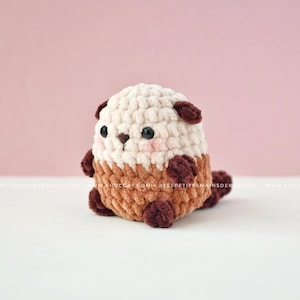 K&ouml;nnte beinhalten: Ein handgefertigtes H&auml;keltier-Otter-Pl&uuml;schtier in Creme, Braun und Dunkelbraun. Das Pl&uuml;schtier hat einen abgerundeten K&ouml;rper, kleine dunkle Augen und rosafarbene Wangen. Der K&ouml;rper des Otters besteht aus einer Mischung aus strukturierten H&auml;kelstichen.