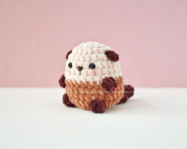 K&ouml;nnte beinhalten: Ein handgefertigtes H&auml;keltier-Otter-Pl&uuml;schtier in Creme, Braun und Dunkelbraun. Das Pl&uuml;schtier hat einen abgerundeten K&ouml;rper, kleine dunkle Augen und rosafarbene Wangen. Der K&ouml;rper des Otters besteht aus einer Mischung aus strukturierten H&auml;kelstichen.