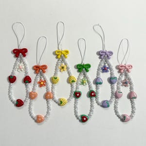 May include: Six phone charms featuring white pearl-like beads, each topped with a colored plastic bow. The charms include various fruit-shaped beads such as strawberries, oranges, and peaches, along with small flower accents in matching colors.