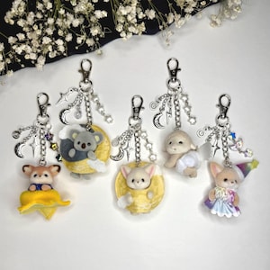 May include: Five keychains featuring small flocked animal figures. One is a koala on a yellow moon, one a fox in a yellow circle, one a lamb, one a fox in a yellow flower, and one a fox wearing a rainbow hat and dress. Each has a silver lobster clasp.